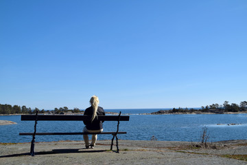 Blonde young woman, in her 30s, all alone, sitting on a bench, enjoying the view of the archipelago...