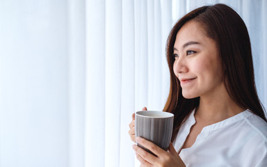 A beautiful asian woman drinking hot coffee while looking outside the window in the morning