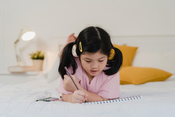 Young Asian girl drawing at home. Asia japanese woman child kid relax rest fun happy draw cartoon in sketchbook before sleep lying on bed, feel comfort and calm in bedroom at night concept.