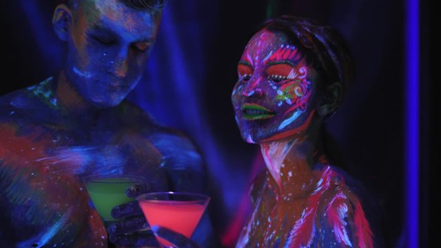 A couple with UV body art dancing with glasses of colorful glowing cocktails in the dark under the light of fluorescent lamps. Art design of couple disco dancers posing in UV.