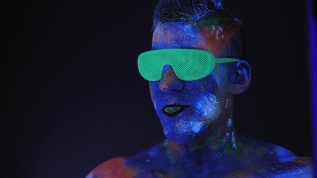 A man with UV drawings on his face and green neon glasses inflates a bubble of bubblegum in the dark under fluorescent lighting. Bright fluorescent body art glows in darkness. UV paint.