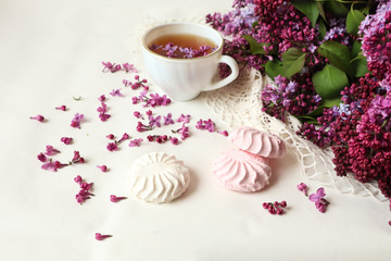 Multi-colored marshmallow with scattered lilac flowers and a white Cup of tea on a white background with a bouquet of lilac, a place for the inscription, the concept of a good morning.
