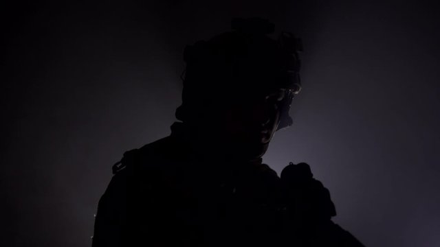 A military soldier in modern outfit with weapons is standing in the smoke in the studio. Military conflict concept, back light, shadow. Slow motion