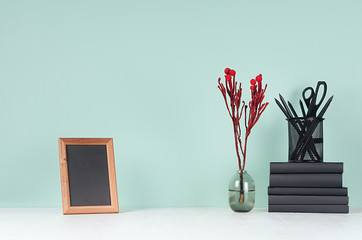 Home workplace with black stationery, books, red branch in glass vase, blank wood photo frame in trend green mint menthe color interior and white wood table, copy space.