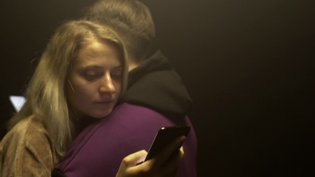 Beautiful couple in love guy and girl stand embracing and look at smartphones on a black background. Hostages of new technologies and smartphones, copy space. Slow motion