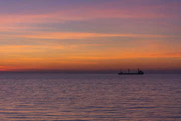 Fototapeta na wymiar Bright twilight sunset sky over the sea. Wonderful sky after sunset in orange shades. The natural background. Purple hue of the sea and sky. Abstract lines of fire clouds. A ship on the horizon.