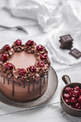 Cherry chocolate cake ornate with cream, cherries and dripping chocolate on grey plate, small bowl with cherries, chocolate bar pieces, napkin and fork on white wooden background. Vertical orientation