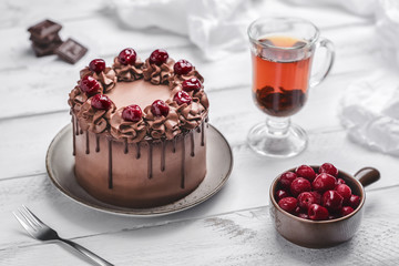 Cherry chocolate cake ornate with cream, cherries and dripping chocolate on grey plate, glass teacup, small bowl with cherries, chocolate bar pieces, napkin and fork on white wooden background
