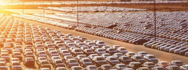 New cars parked in a distribution center on a sunset or sunrise and car factory buildings with pipes. Parking in the open air.