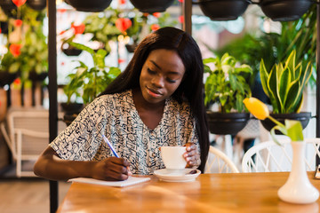 Portrait of beautiful young african woman sitting at cafe and writing notes