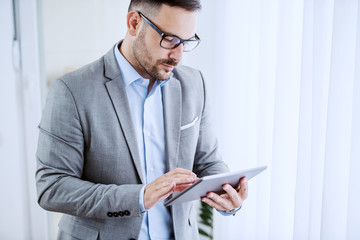 Serious caucasian classy businessman in suit and with eyeglasses standing next to window and using tablet at his office.