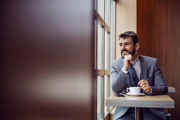 Young attractive businessman sitting in cafeteria, stirring coffee and looking trough window. He is on coffee break.