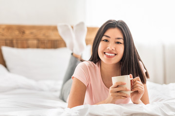 Asian Girl Having Coffee Lying In Bed In Cozy Bedroom
