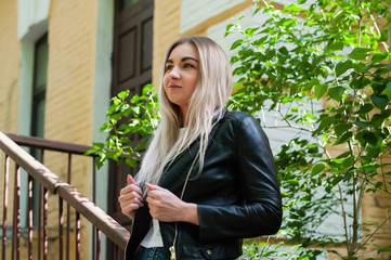 Nice blonde girl in a black leather jacket in spring on background of green leaves, trees, near a beautiful building