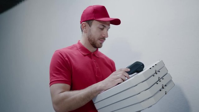 Young delivery man in red uniform delivering pizza to a customer's house. Beautiful female client receiving fast food paying for her order with paypass smartphone application. NFC technology.