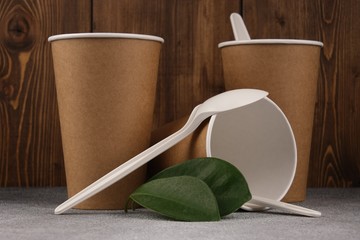 Three cardboard cups with green leaves lie on a concrete surface on a wooden background, in one glass an eco-friendly spoon. Mock-up. Close-up.