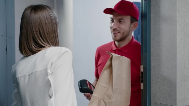 Young woman opens door to delivery man receiving a package with fresh heallthy food paying with paypass technology smartphone application. Online order, home delivery.