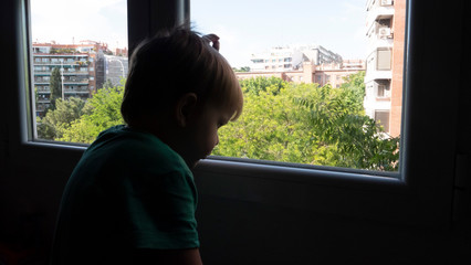 Caucasian blond boy of 3 years looks out the window of his house in a melancholic way