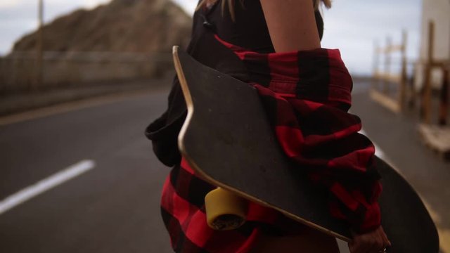 Cropped footage woman in plaid coat on a cloudy day at walking, holding a skateboard, slow motion. Unrecognizable girl walks by asphalt road