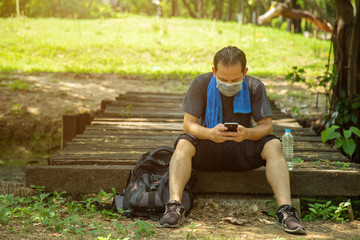 Fototapeta premium Asian man in black casual wear wearing a face mask sitting on old boards of a wooden bridge using an application on cell phone in public park with grreen lawn nature background