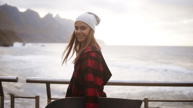 Close up footage sporty woman in hat, plaid coat enjoying time by the seaside on a cloudy day at sunset, walking freerly, holding a skateboard. Wind fluttering her hair, posing, smiling for camera