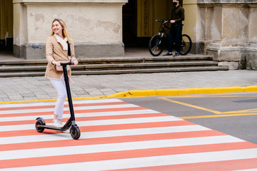 Female courier with yellow backpack on an electric scooter. Fast delivery in city street over blooming tree at spring time.