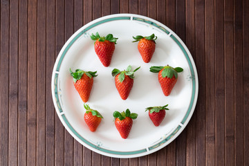 Many strawberries arranged in a white ceramic plate with green decoration on the edge, the composition is resting on a wooden table.