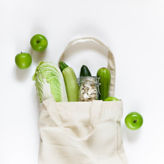 Reusable canvas bag with green apples, pumpkin seeds in a glass jar, fresh vegetables on a white background. Concept zero waste shopping, healthy eating, vegetable food. Flat lay, top view, close-up.