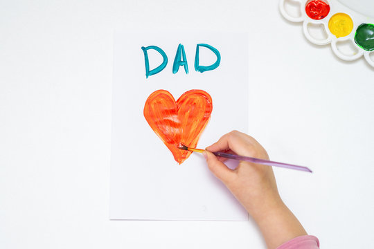 Top view of child's hand drawing red heart with word Dad greeting card on white paper. Happy Father's Day concept.