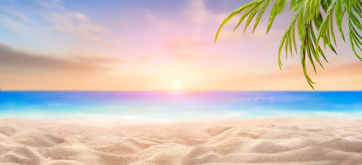 A summer vacation, holiday background of a tropical beach and blue sea at sunset with the sun on the horizon and green palm tree leaves.