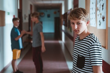 Somber boy is standing in the school corridor. Behind him are two classmates