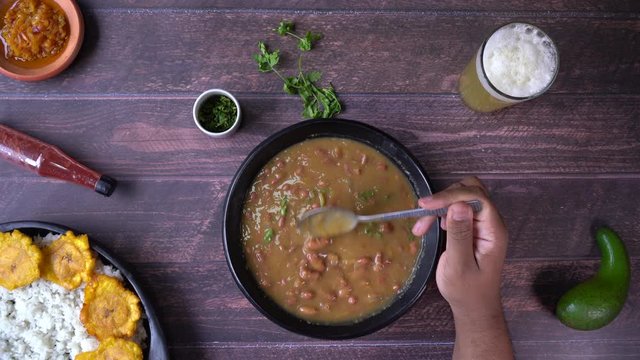 Above view of a black man&rsquo;s hand stirring bean curry in a rustic artisanal bowl on a set table including a rice dish with fried plantain and fresh naranjilla juice