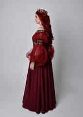 Portrait of a beautiful woman with red hair wearing  a  flowing Burgundy fantasy gown and golden crown.  full length standing pose, isolated against a studio background
