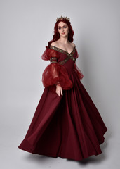 Portrait of a beautiful woman with red hair wearing  a  flowing Burgundy fantasy gown and golden crown.  full length standing pose, isolated against a studio background  © faestock