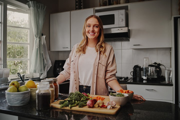 Young charming woman posing in kitchen standing near table with fresh organic vegetables and...