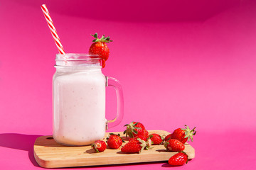 Strawberry smoothie in a glass jar with a red-striped tube. There are strawberries on a wooden Board and a strawberry smoothie next to them. Concept of healthy food. On a bright pink background