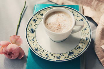 Cup of hot coffee with milk on plate, old book and rose flower on bed linen, coffee in bed. Copy space flat lay