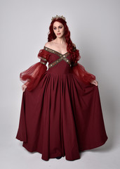Fototapeta premium Portrait of a beautiful woman with red hair wearing a flowing Burgundy fantasy gown and golden crown. full length standing pose, isolated against a studio background 