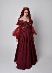 Fototapeta premium Portrait of a beautiful woman with red hair wearing a flowing Burgundy fantasy gown and golden crown. full length standing pose, isolated against a studio background 