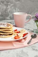 A stack of pancakes with a pink tea mug and  fresh strawberries on a white plate on a light background. A small bouquet of lilacs in a glass vase stands next to the Breakfast.