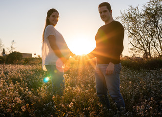 Young pregnant couple holding hands into white flowers field with the sunset and sun rays in the background