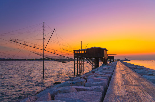 Traditional fishing station house with fishnet in water of Adriatic sea at pier Diga Sottomarina, skyline with beach, amazing yellow red sunset at twilight, dusk, evening view, Northern Italy