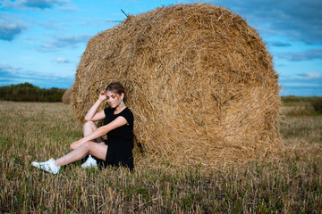 Naklejka na ściany i meble Style fashion on street. A girl with bundle hair in a earrings, black dress and white sneakers. Female portrait. Model posing. A girl in a dress on a haystack background. Fashion photography