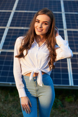 Pretty woman with with long hair smiling on background of solar panels at warm day. Girl in jeans and a white shirt. Solar energy concept image