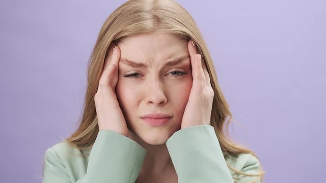 A close-up view of a tired stressful young woman is feeling a headache standing isolated over gray background