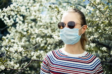 Young girl in medical mask and sunglasses on background of blossoming apple tree on summer day.