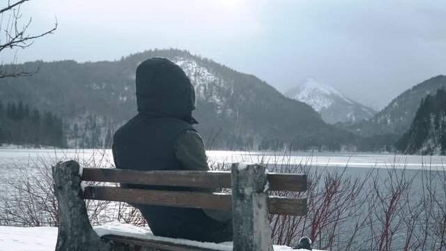 The man in the hood sits on a bench with his back by the lake in winter and looks at the mountains.