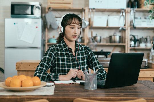 portrait of asian young lady with earphones taking online language course in dining room. Chinese girl sitting straight is repeating new words after teacher. study from home concept