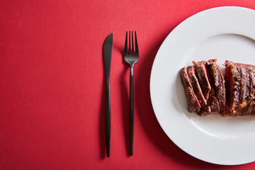 top view of tasty grilled steak served on plate with cutlery on red background