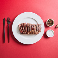 top view of tasty grilled steak served on plate with cutlery and salt and pepper in bowls on red background